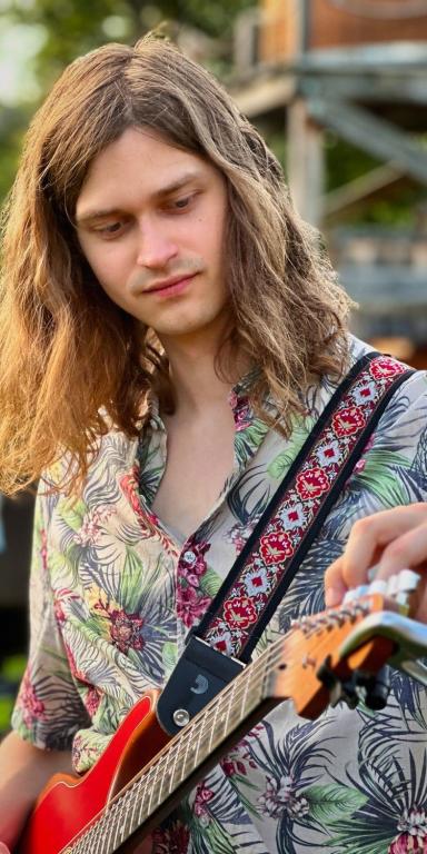 Gitarrist Anton im Porträt stimmt seine Gitarre, er trägt lange Haare und hat ein Hemd mit Blumenmustern an