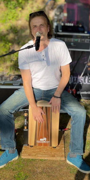 Dirk sitzt auf seinem Cajon, das hellbraun ist und mit einem holzfarbenen Streifen versehen, er trägt Jeans und ein weißes Shirt