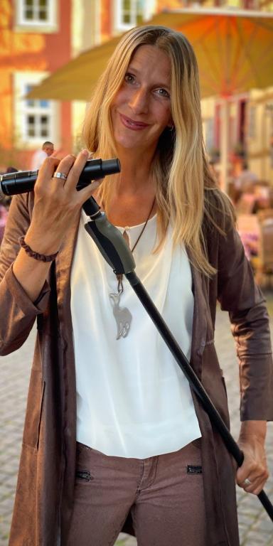 Sängerin Stephanie steht mit ihrem Mikrofon im Mikrofonständer in der Hand auf einem Platz, sie hat lange, blonde Haare, trägt ein weißes Shirt und darüber eine braune, längere Jacke