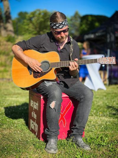 Gitarrist Toni sitzt auf einem roten Sitzwürfel auf einer Wiese und spielt Akustikgitarre, er trägt eine Jeans mit Rissen und hat um den Kopf ein Bandana gebunden, darunter einen Pferdeschwanz